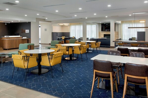 A hotel lobby with multiple seating areas, including round tables and chairs, a reception desk, and a kitchenette in the background. - Courtyard by Marriott New Haven Orange-Milford (Orange)