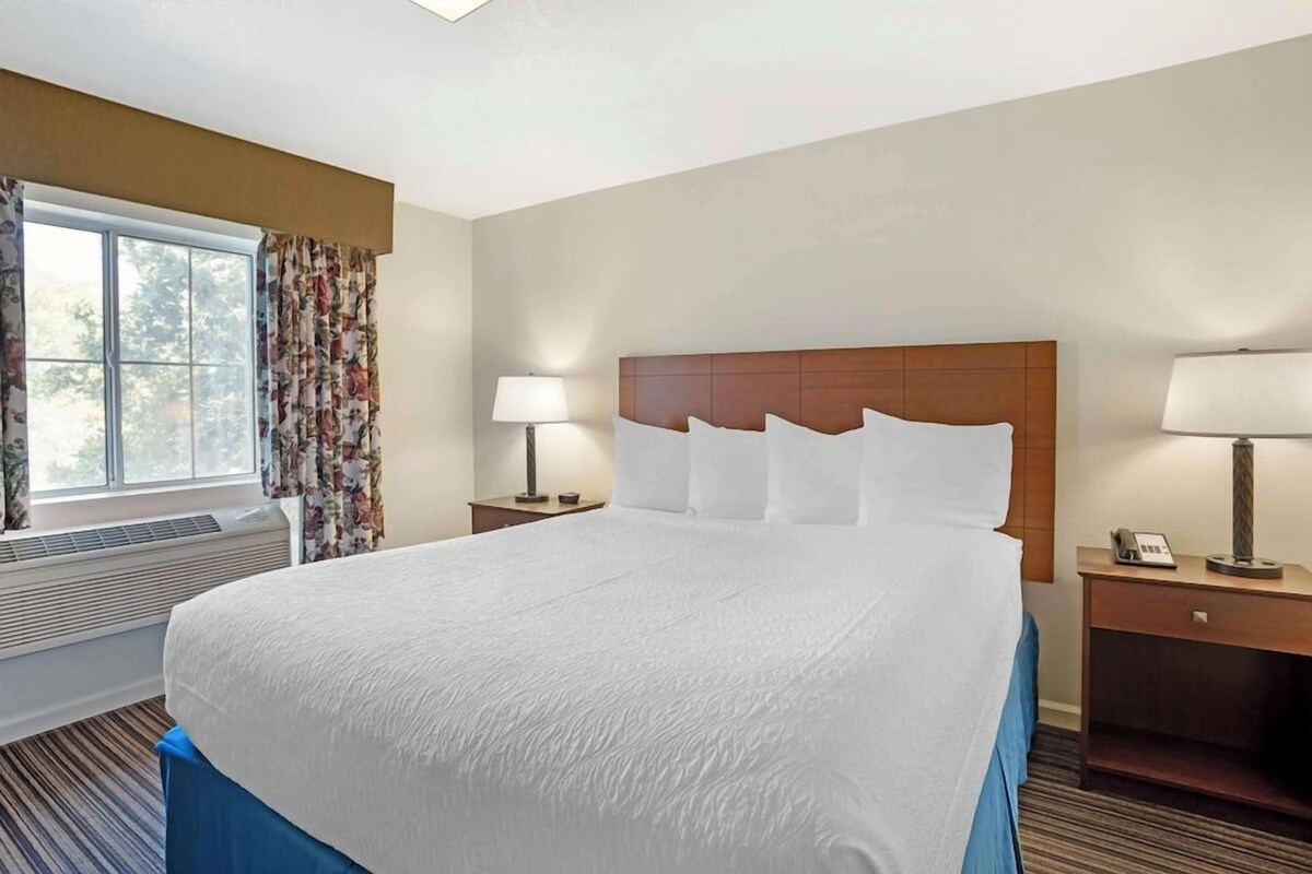 A hotel room with a large bed, two bedside tables with lamps, a window with floral curtains, and a view of trees outside.