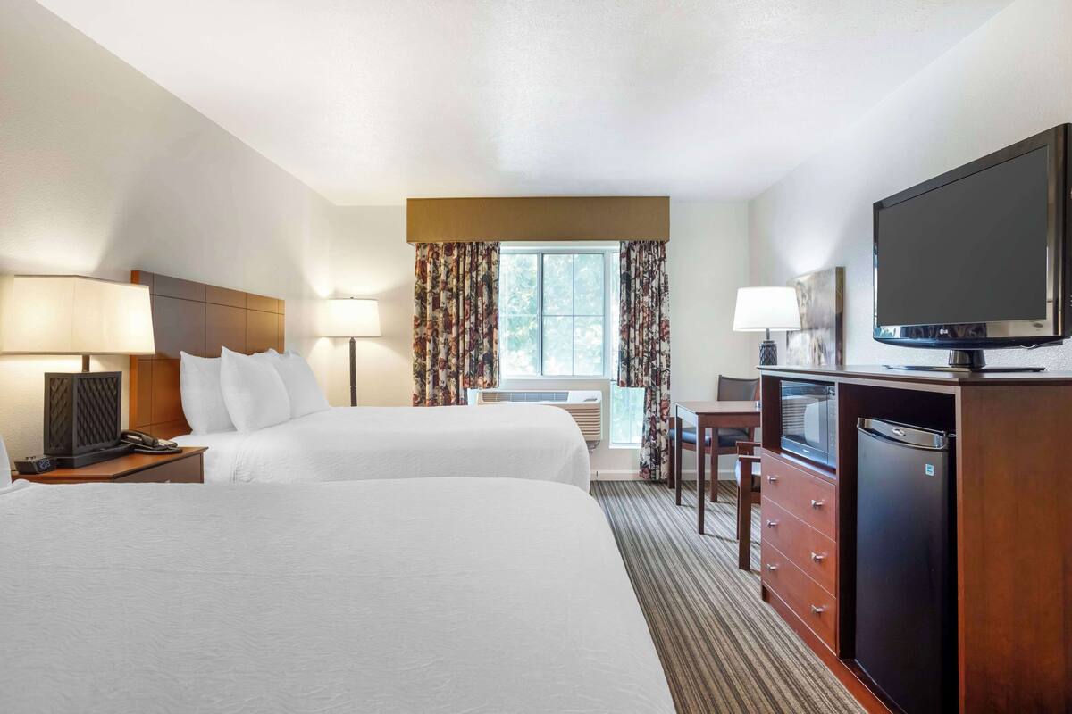 A hotel room with two beds, a desk with a television, a refrigerator, and a window with curtains.