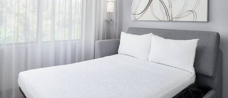 Uma cama bem arrumada com colcha branca, cabeceira estofada cinza e uma obra de arte abstrata na parede acima.