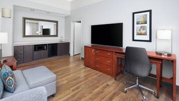 A hotel room with a wooden floor, a grey sofa, a desk with a chair, a TV, a mirror, and a framed picture on the wall.