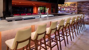 A bar area with a long counter, multiple bar stools, and a well-stocked shelf of bottles. - Courtyard by Marriott Scottsdale North (Phoenix)