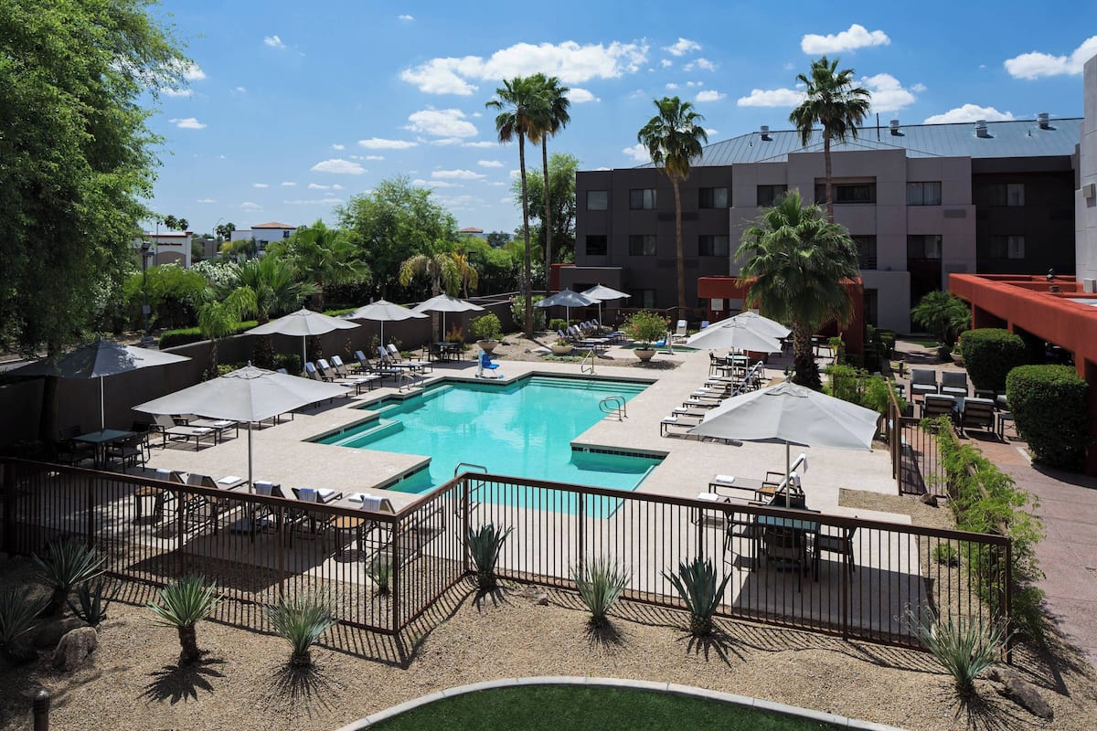 A hotel pool area with a rectangular pool, lounge chairs, umbrellas, and palm trees.