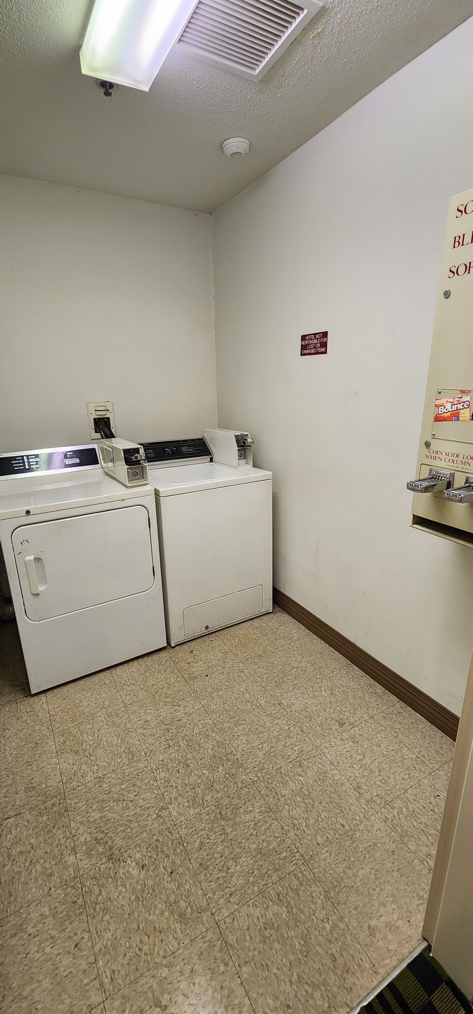 A laundry room with two white washing machines, a ceiling vent, a smoke detector, and a sign indicating the cost of coin-operated laundry services.