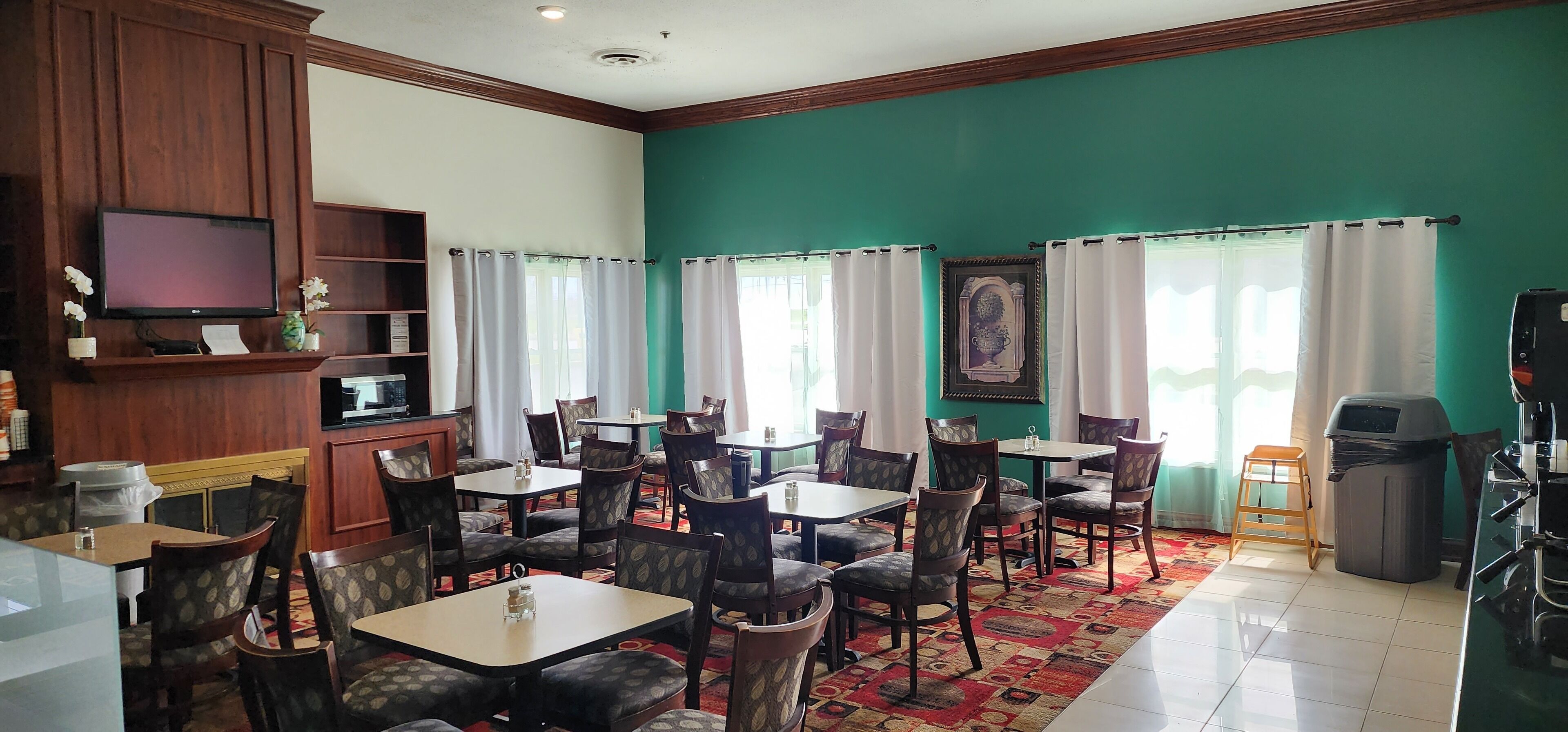Um restaurante com painéis de madeira, paredes verdes e cortinas brancas. Mesas e cadeiras dispostas para refeições.