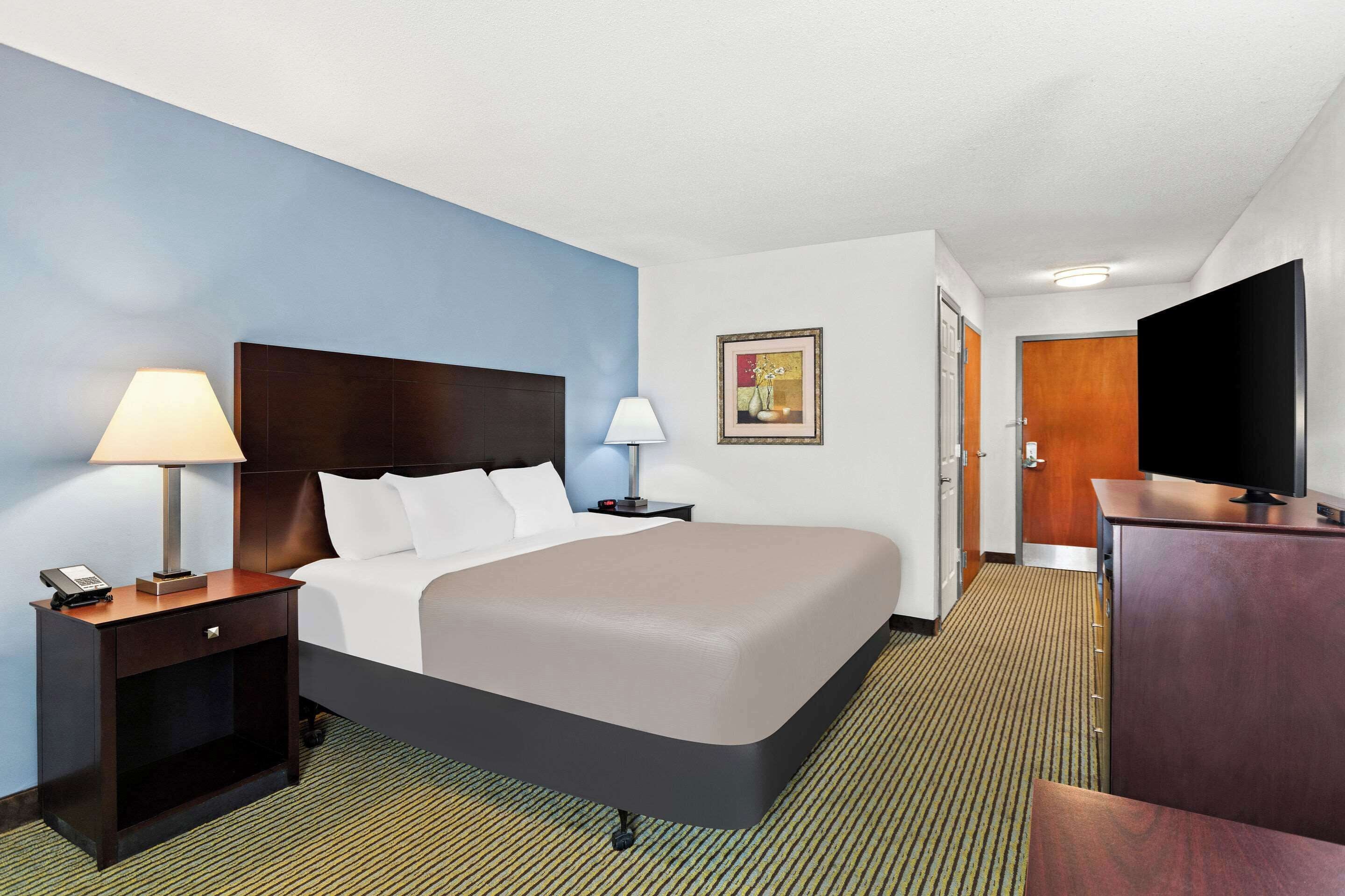 A hotel room with a large bed, a nightstand with a lamp, a desk with a TV, and a door leading to another room.