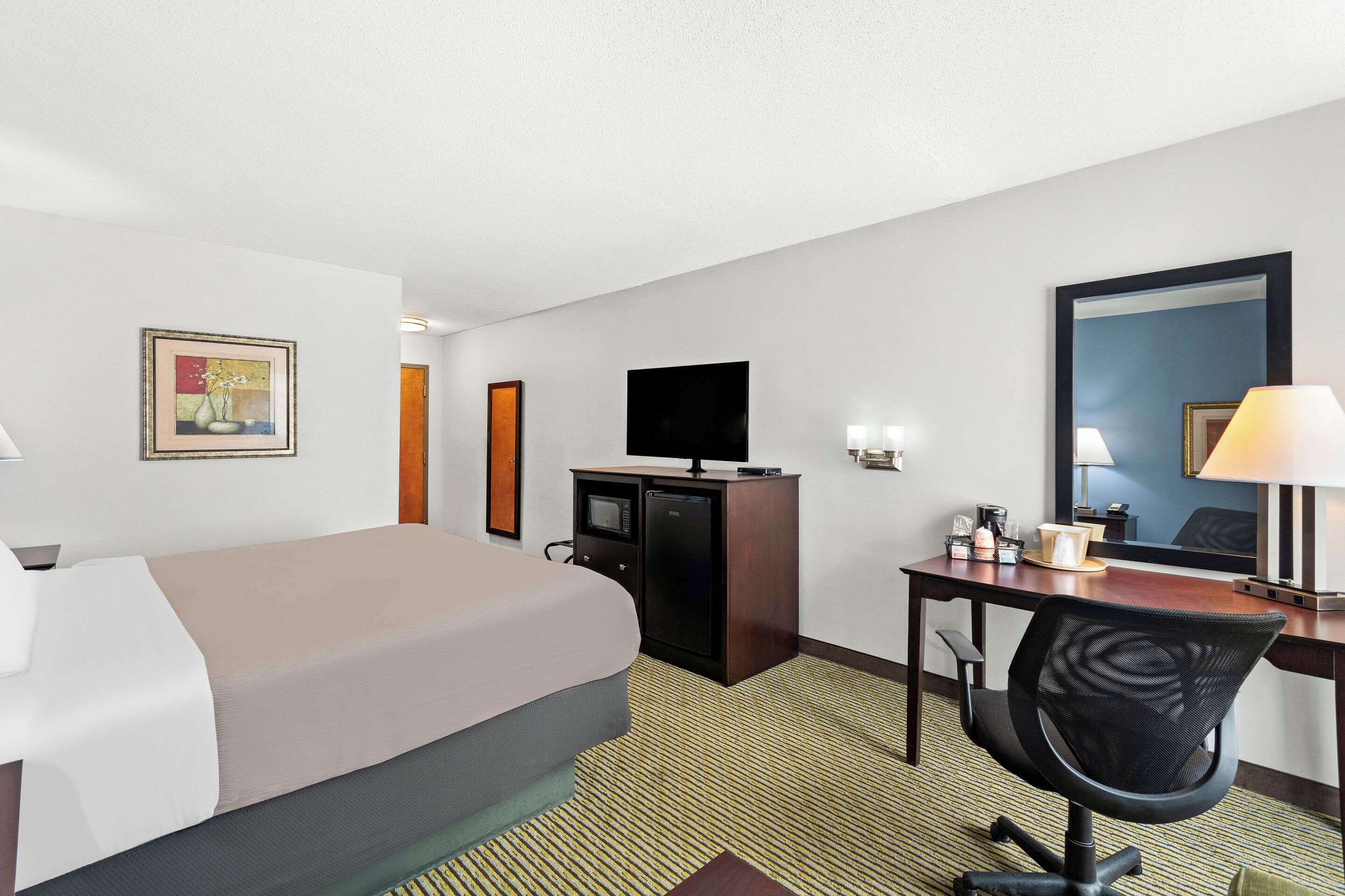 A hotel room with a bed, a desk with a chair, a television, a mirror, and a painting on the wall.
