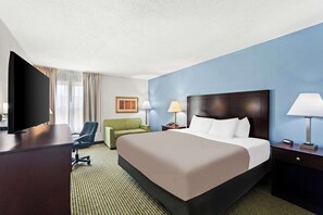 A hotel room with a large bed, a desk with a flat-screen TV, a green armchair, two bedside lamps, and a framed picture on the wall.