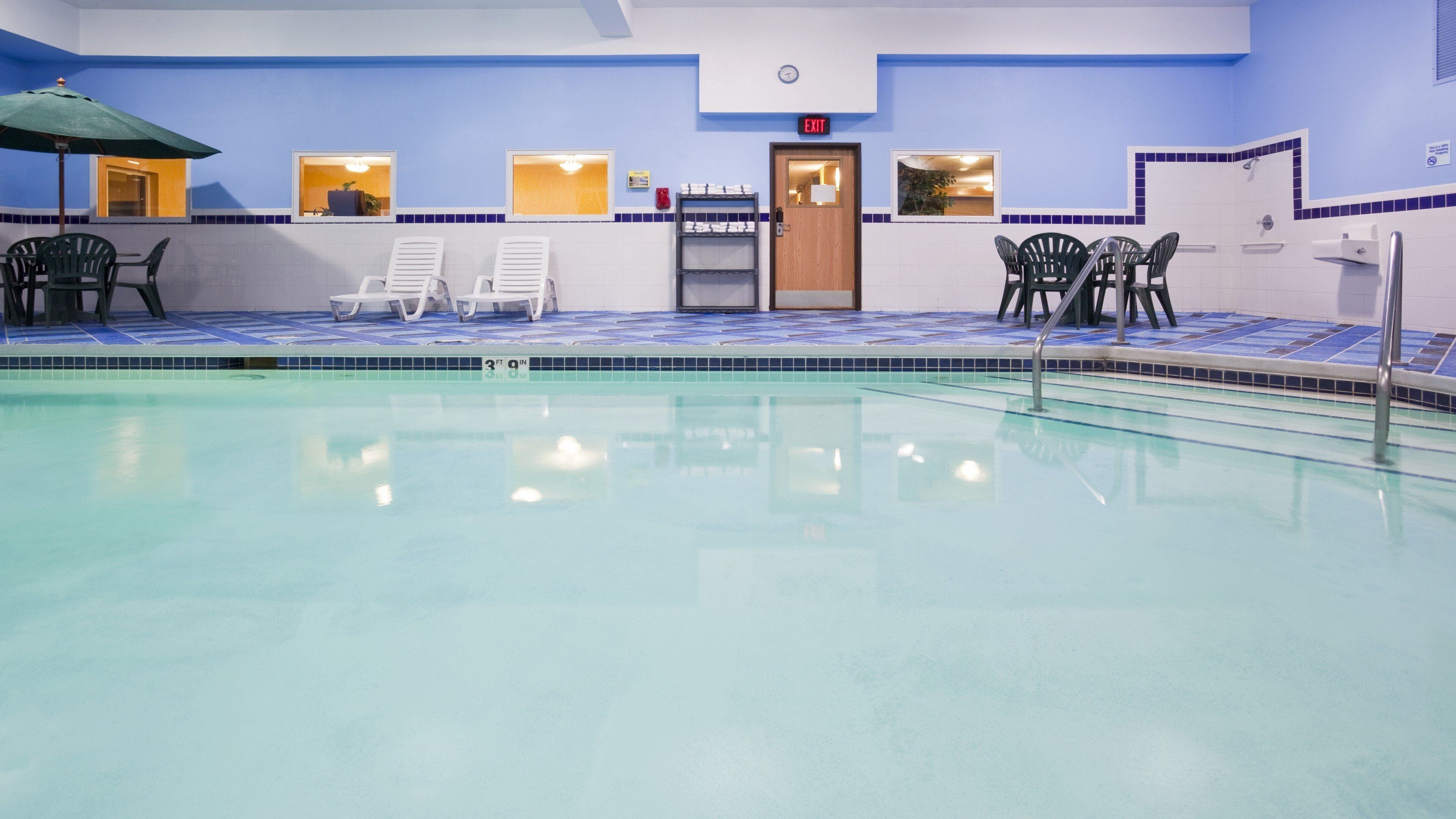 An indoor swimming pool with clear water, surrounded by lounge chairs and a reception area with an exit sign.