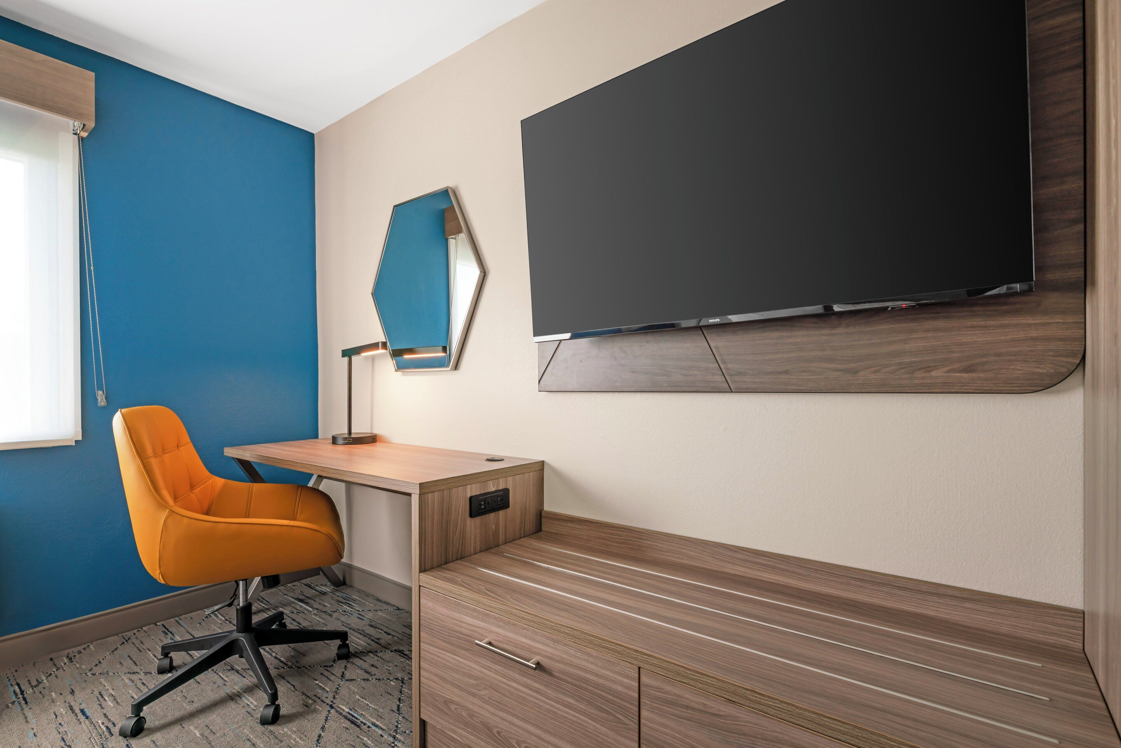 A room with a desk, an orange chair, a flat-screen TV, and a wall-mounted mirror.