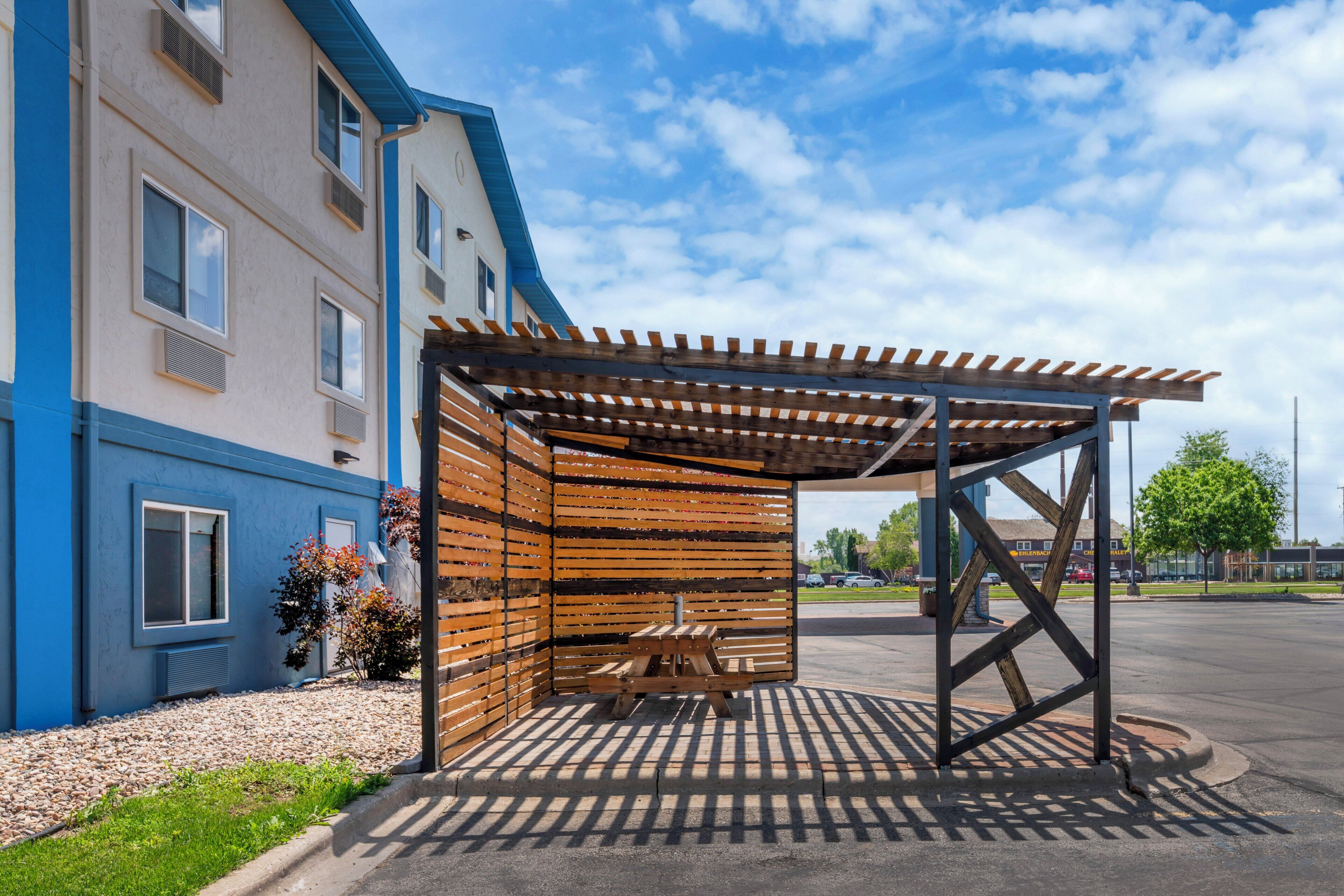 Un espace extérieur aménagé, abrité et doté d’une pergola en bois, jouxtant un bâtiment avec de nombreuses fenêtres et une façade bleue.