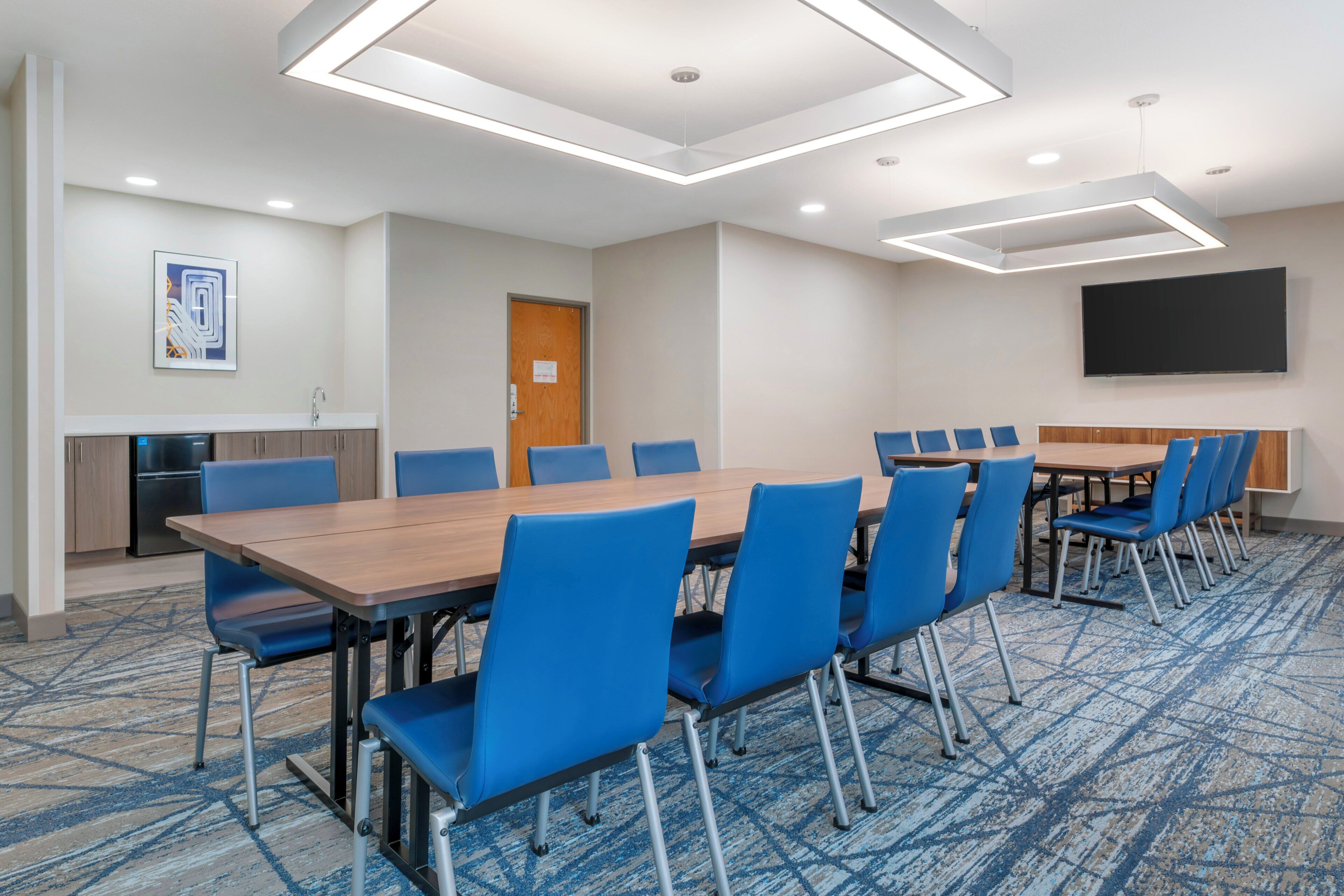 A conference room with a long table, blue chairs, a flat-screen TV, and a kitchenette.