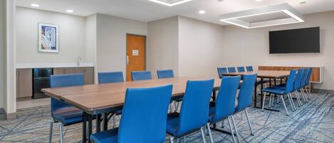 A conference room with a long table, blue chairs, a flat-screen TV, and a kitchenette.