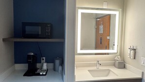 A compact hotel bathroom with a sink, mirror, and a small refrigerator.