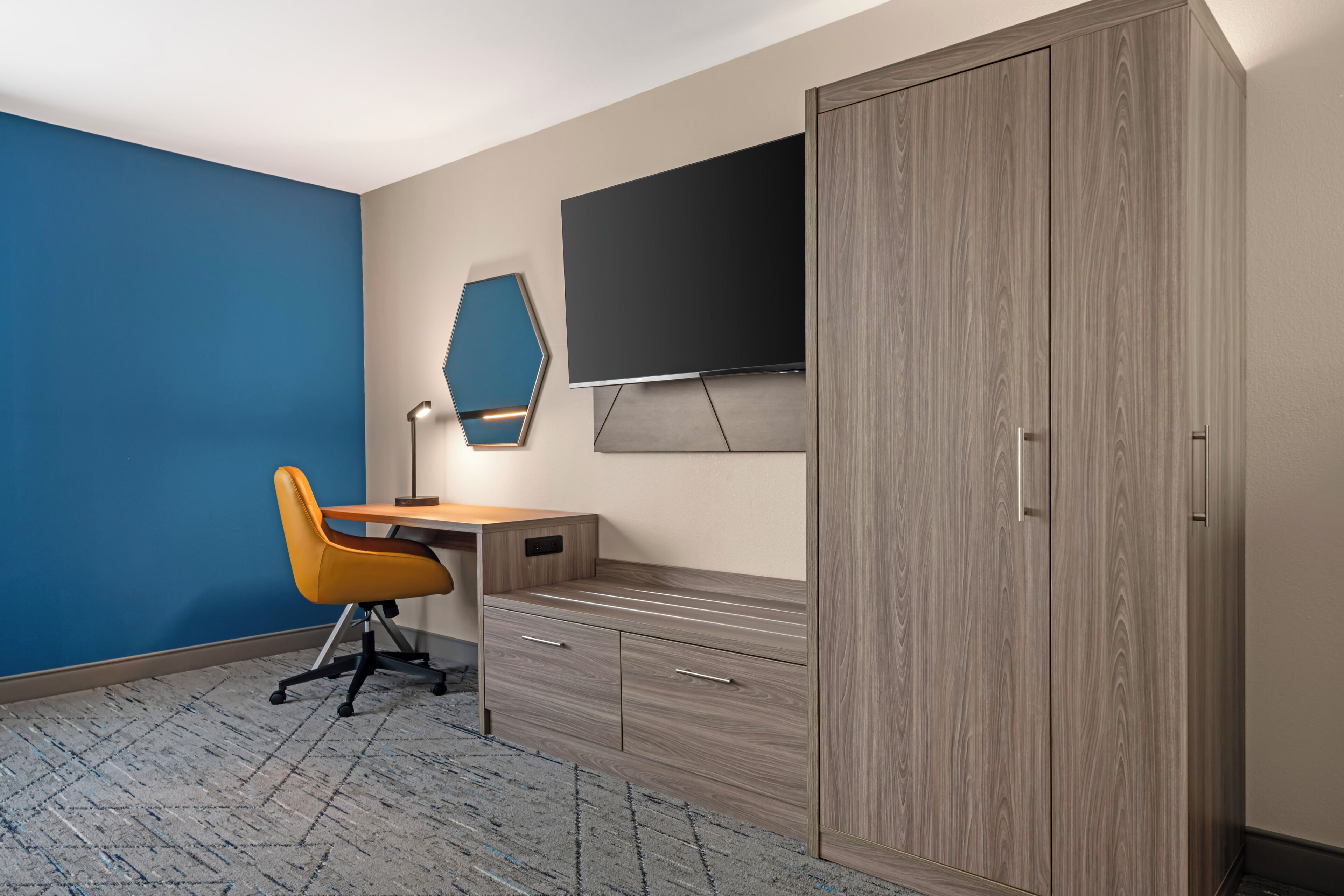 Ein Zimmer mit blauer Wand, einem Holztisch, einem gelben Stuhl, einem Kleiderschrank und einem an der Wand befestigten Flachbildfernseher.