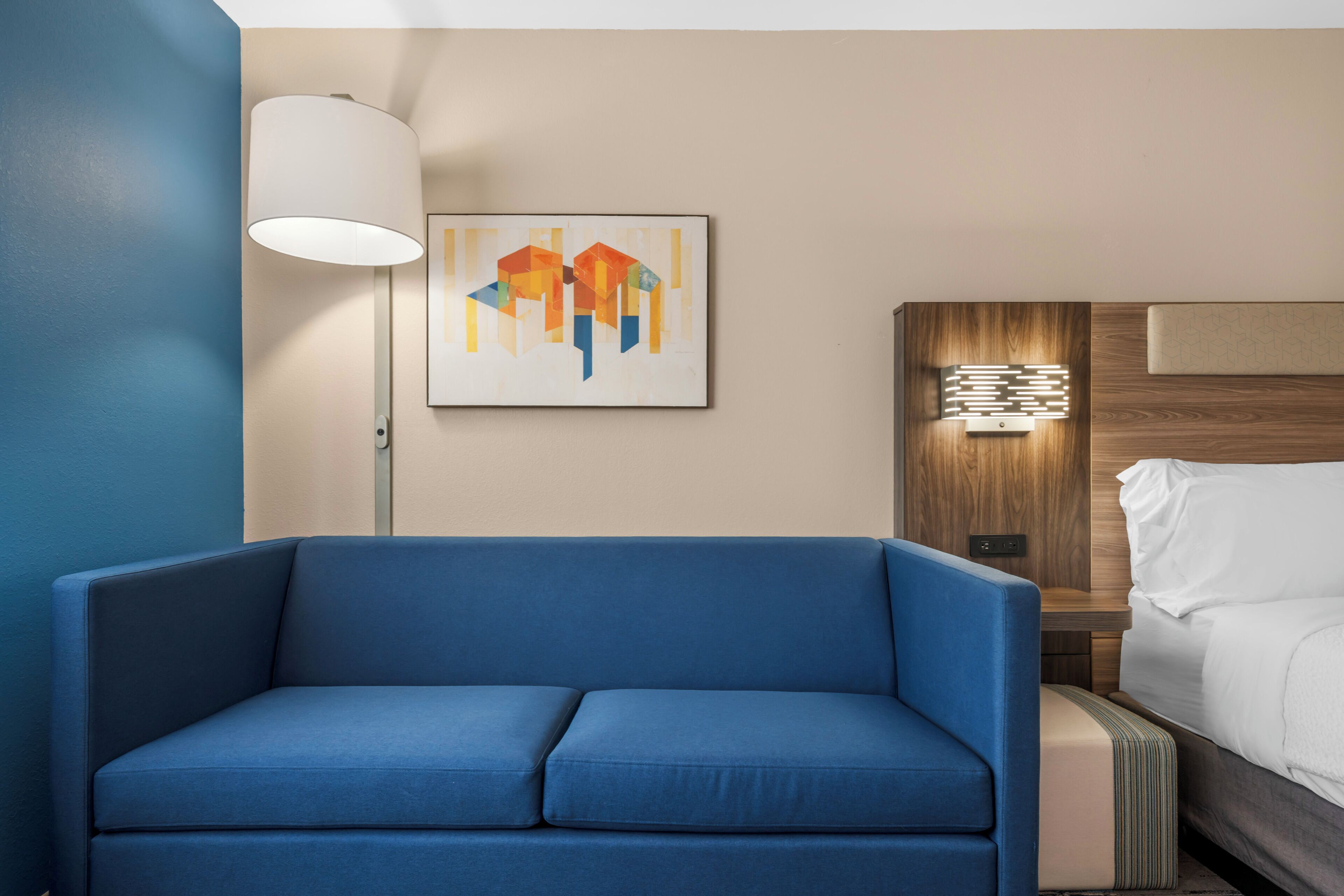 Habitación de hotel con sofá azul, lámpara, una obra de arte en la pared y una cama con cabecero de madera.