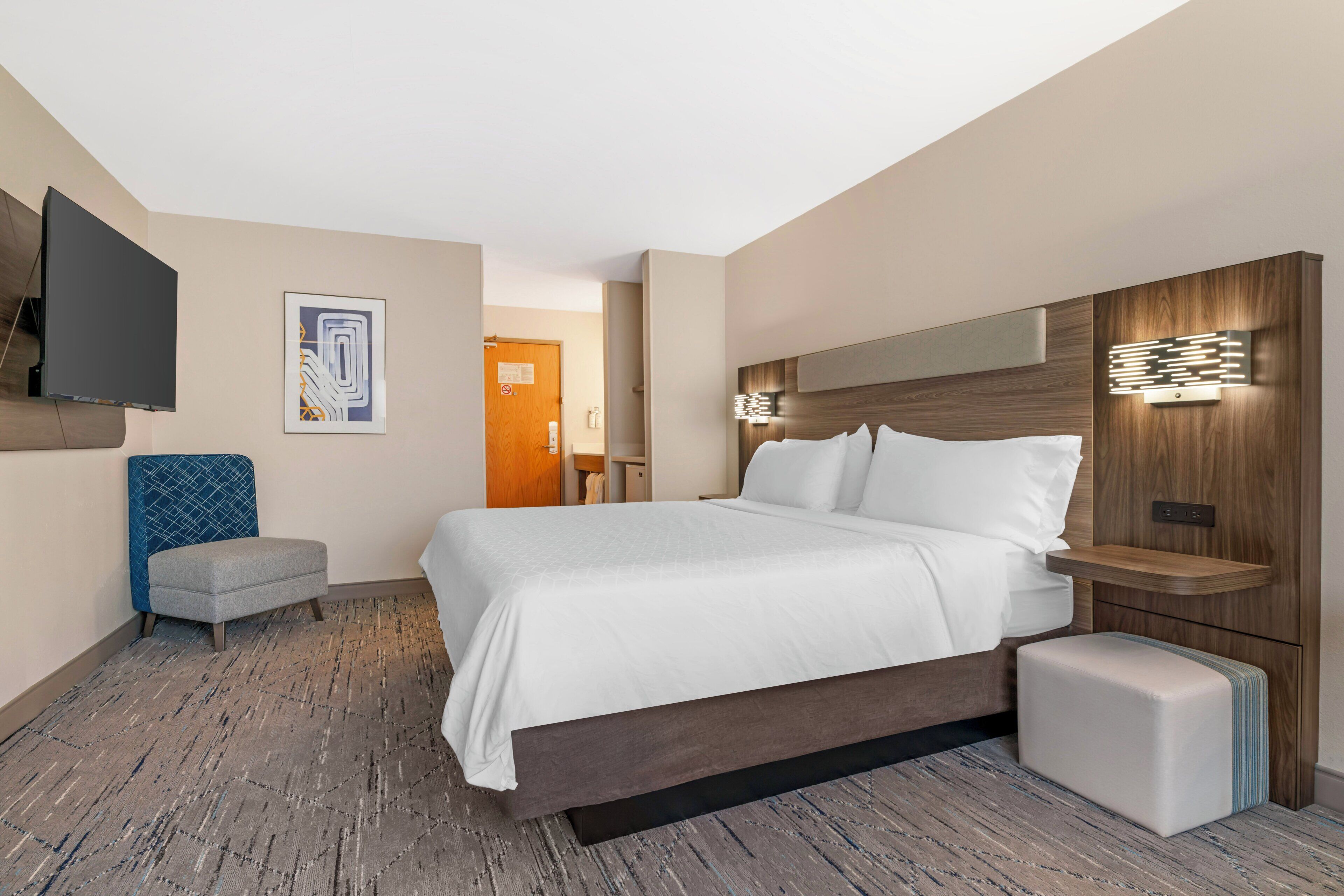 Une chambre d’hôtel dotée d’un grand lit, d’un téléviseur à écran plat fixé au mur, d’un petit coin salon et d’une tête de lit en bois avec éclairage intégré.