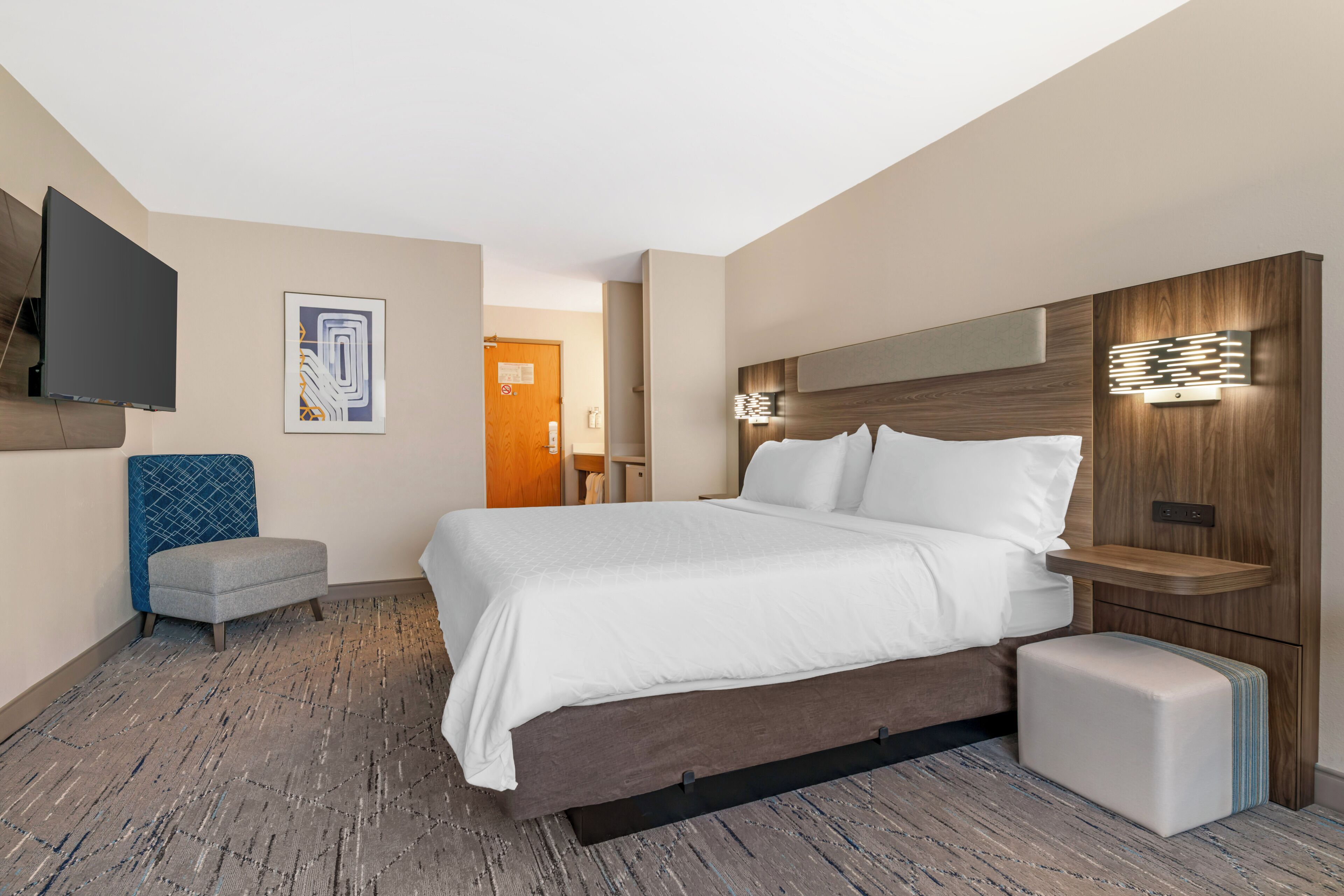 A modern hotel room with a large bed, a flat-screen TV mounted on the wall, a small seating area, and a wooden headboard with integrated lighting.