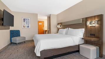 A modern hotel room with a large bed, a flat-screen TV mounted on the wall, a small seating area, and a wooden headboard with integrated lighting.