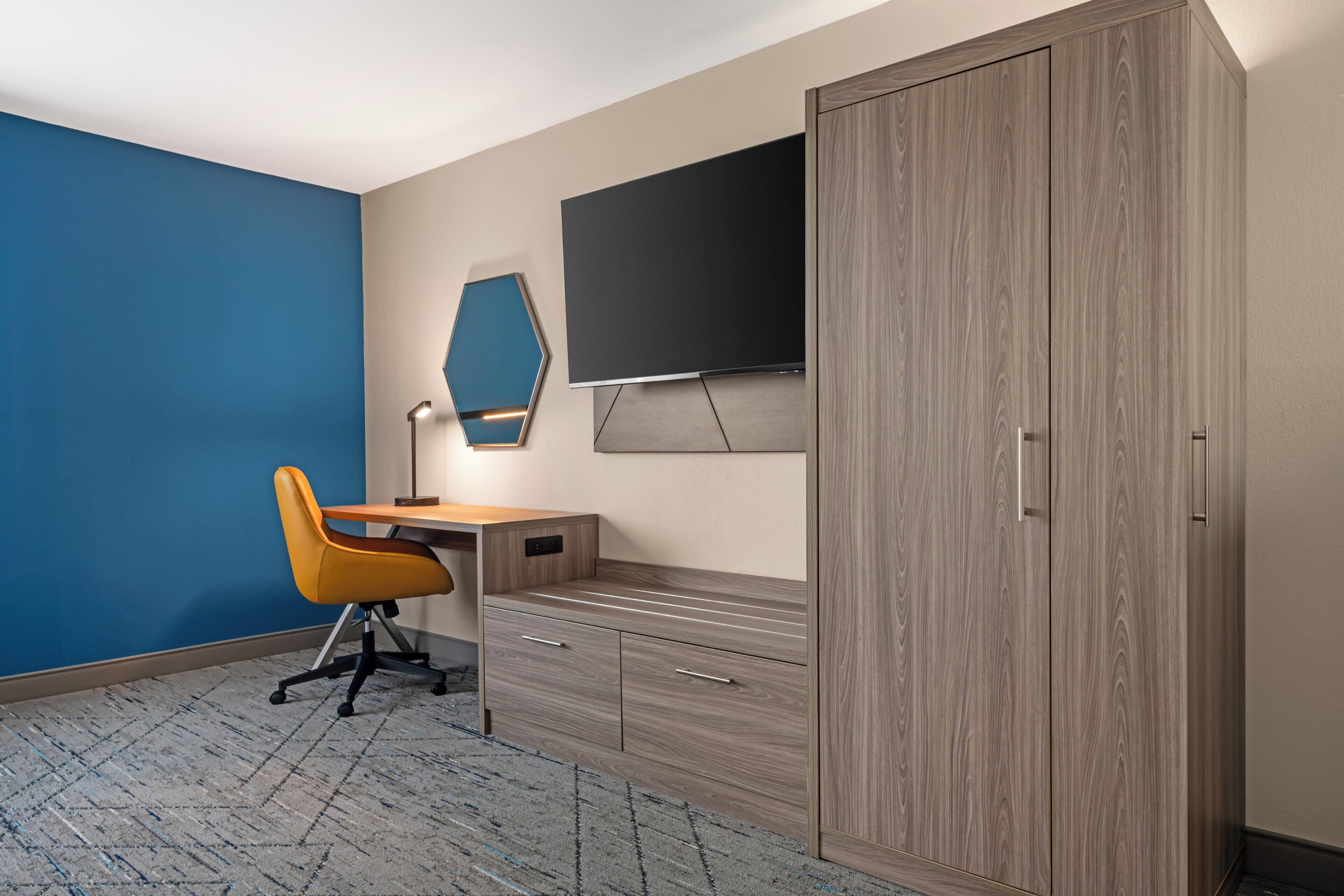 A room with a blue wall, a wooden desk, a yellow chair, a wardrobe, and a flat-screen TV mounted on the wall.