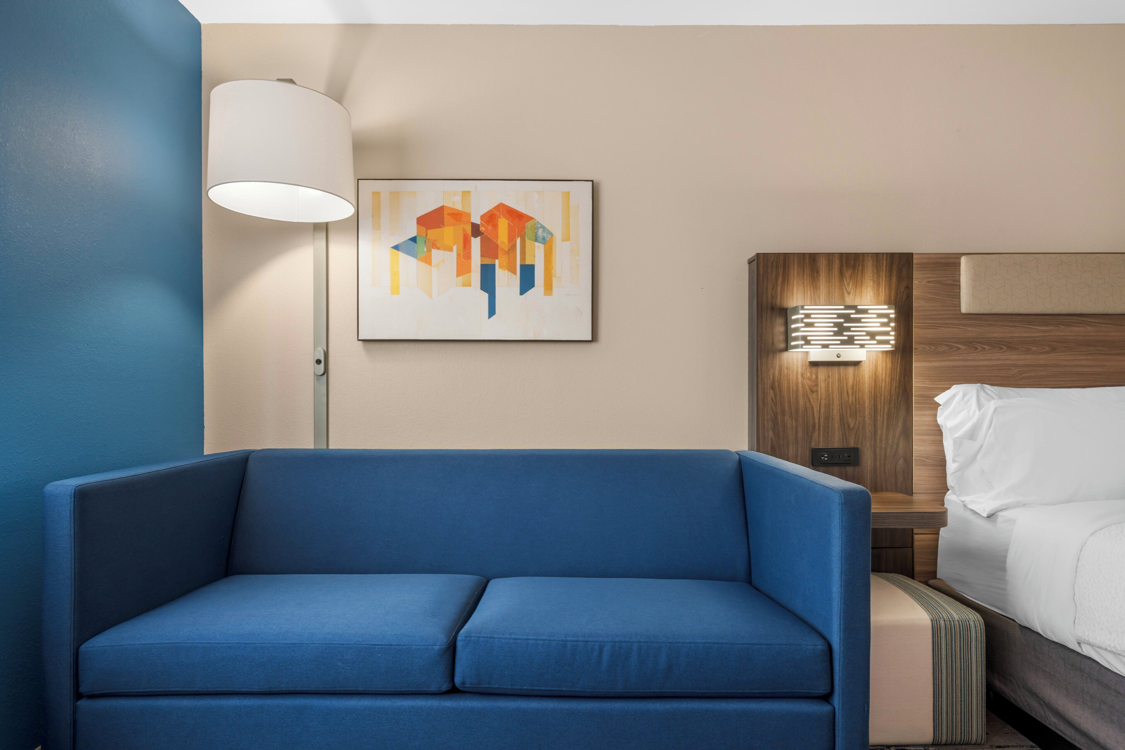 Habitación de hotel con sofá azul, un cuadro en la pared, una mesita de noche con lámpara y cama con ropa blanca.
