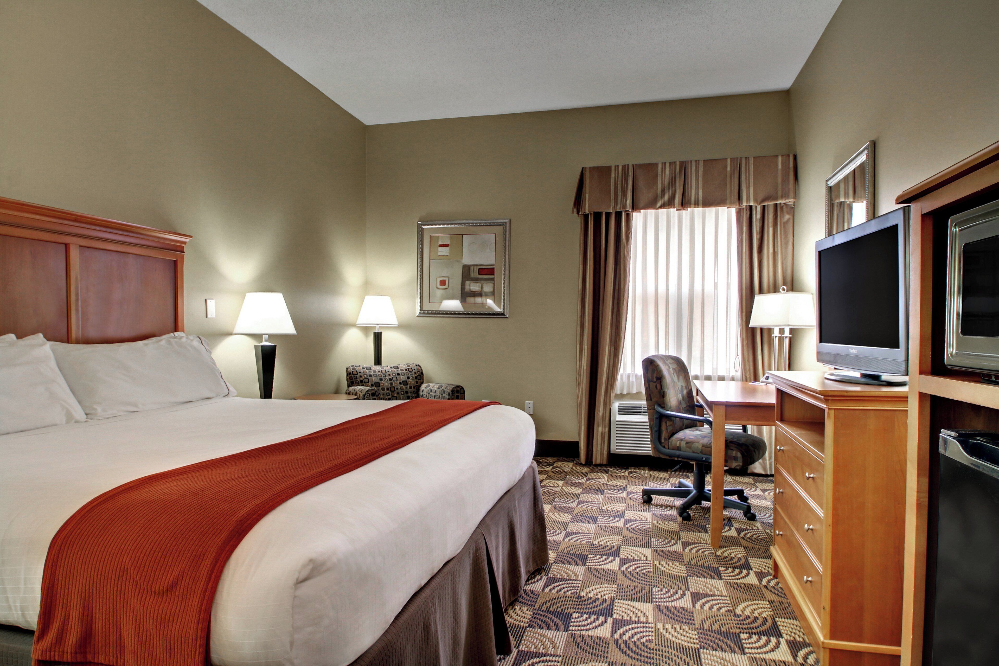 A hotel room with a large bed, a desk with a television, a chair, and a desk lamp.