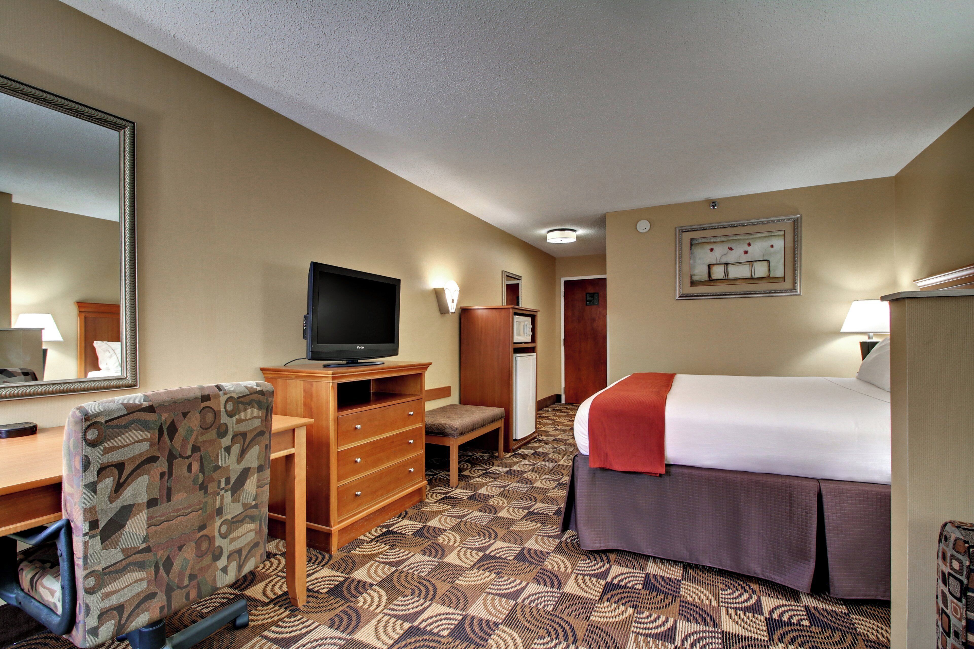 A hotel room with a bed, a television, a desk with a chair, a dresser, and a mirror.