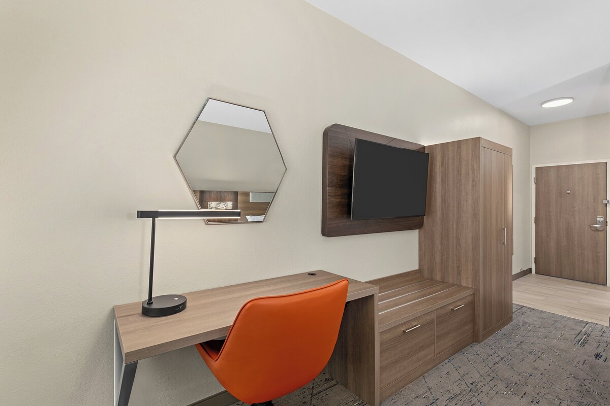 Une chambre d’hôtel avec un bureau en bois, une chaise orange, un grand miroir, un téléviseur à écran plat et une armoire.