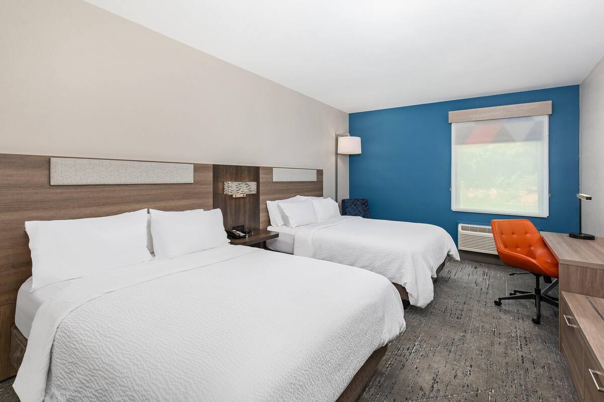 A hotel room with two beds, a desk, an orange chair, a lamp, and a large window.