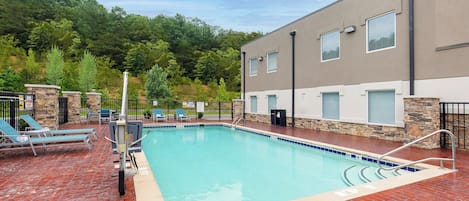 Una piscina rectangular con borde azul, rodeada por una solana de ladrillo rojo. Hay sillas de descanso y un muro de piedra con un pilar de piedra.
