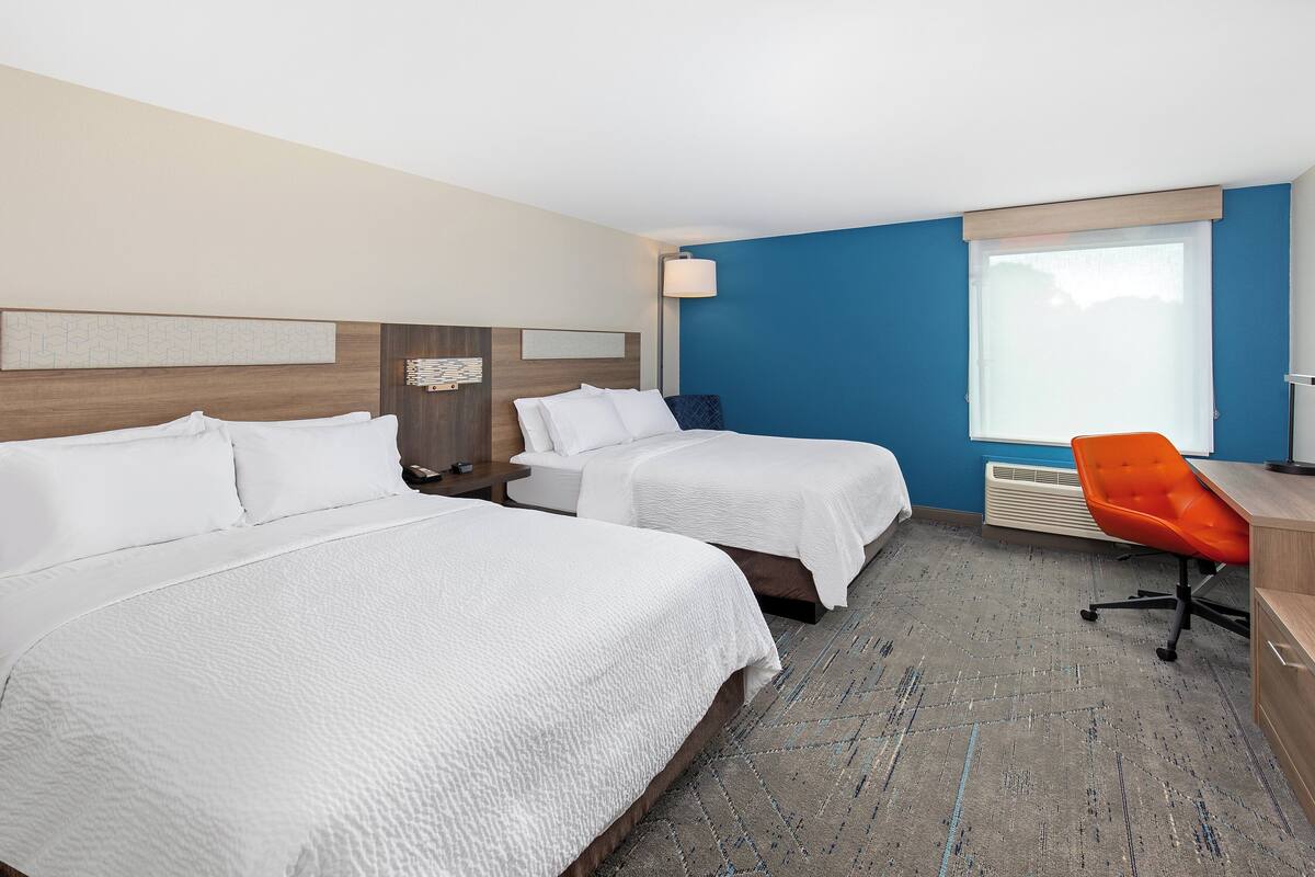 A hotel room with two beds, a desk, an orange chair, and a large window.