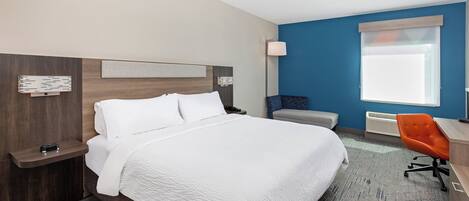 A hotel room with a large bed, a desk with a chair, a window with blinds, and a wall-mounted heater.