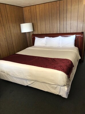 A hotel room with a wooden paneled wall, a bed with white linens and a maroon throw, and a bedside lamp.