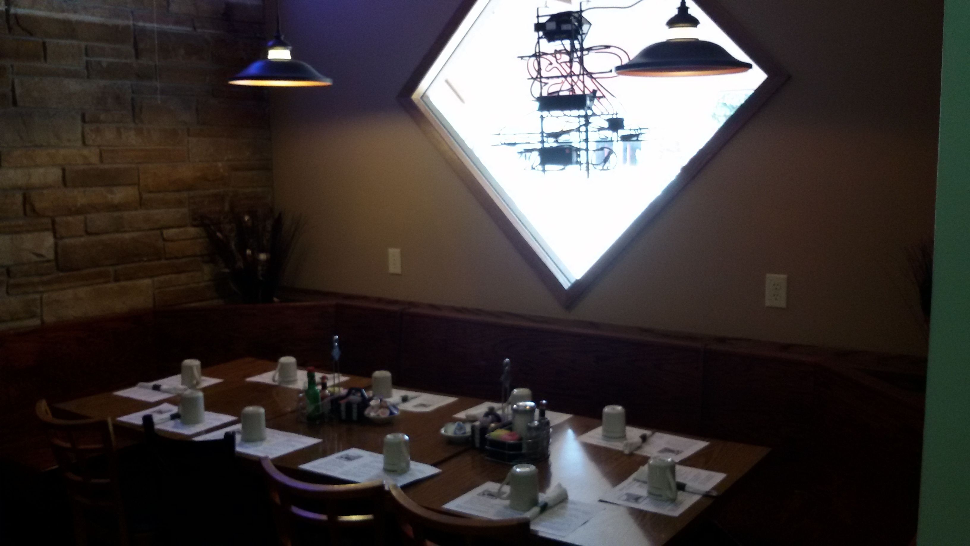 Un restaurante tenuemente iluminado con una pared de ladrillo, un amplio ventanal con diseño geométrico y una mesa de madera preparada para comer.