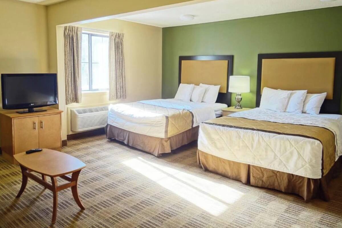 Ein Hotelzimmer mit zwei Betten, einem Fernseher, einem Schreibtisch und einem Fenster mit Vorhängen.