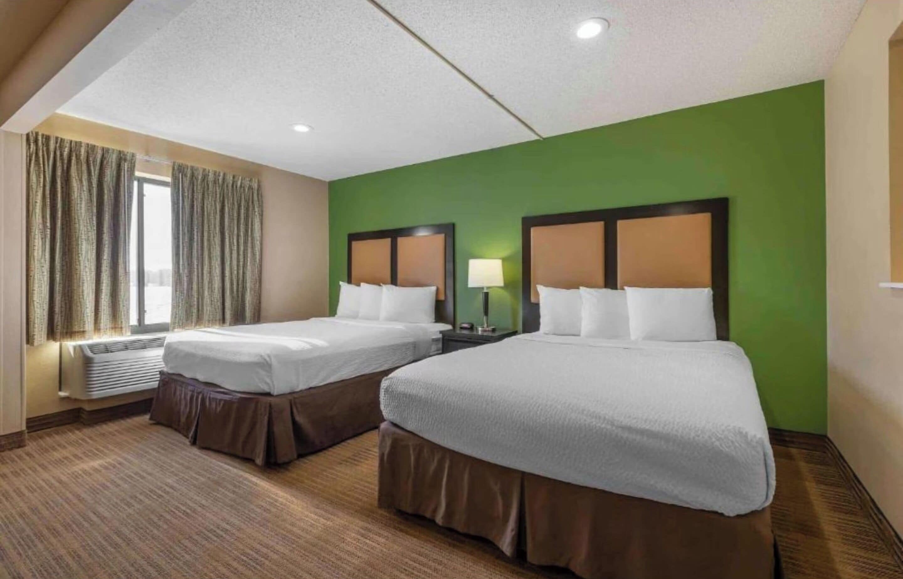 A hotel room with two beds, a window with curtains, a lamp, and a thermostat.