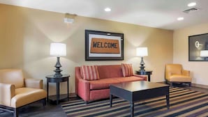 A hotel lobby with a red sofa, two armchairs, a coffee table, and a welcome sign. - Studio 6 Extended Stay South Bend Mishawaka South IN (Mishawaka)