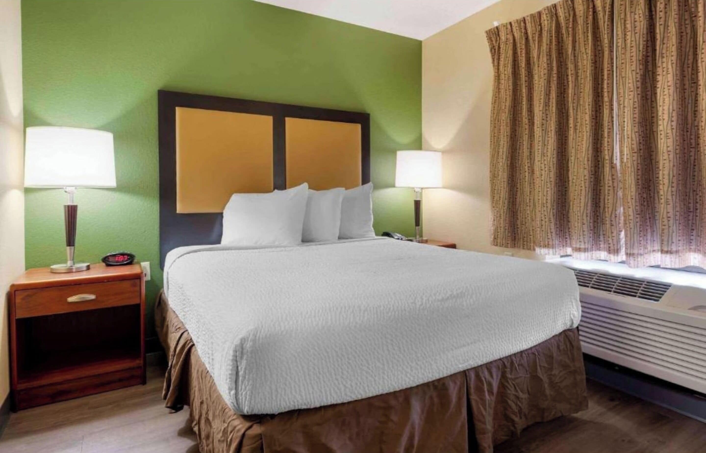 Ein Hotelzimmer mit einem Bett, zwei Nachttischen mit Lampen, einer Wandklimaanlage und einem Vorhangfenster.