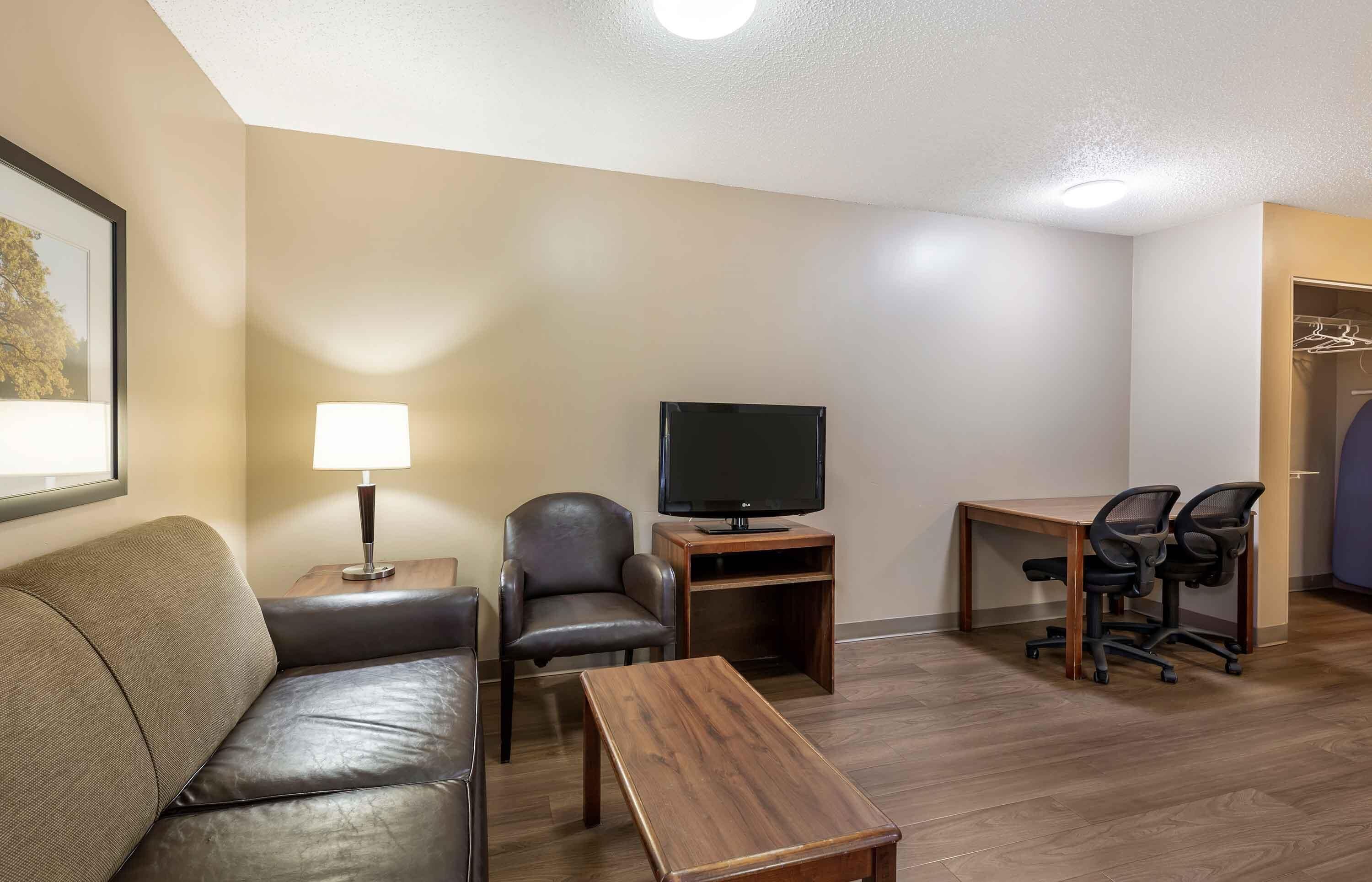 Habitación de hotel con sofá, televisor sobre un mueble de madera, escritorio con dos sillas y armario con ropa colgada.