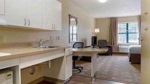 Une chambre d’hôtel équipée d’une kitchenette, d’un bureau, d’une chaise, d’un lit et d’une fenêtre avec des rideaux.