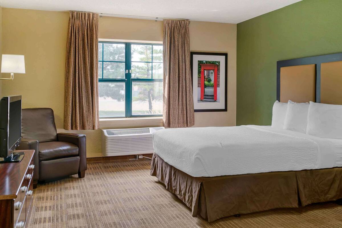 Une chambre d’hôtel avec un grand lit, un bureau avec une chaise, une fenêtre avec des rideaux et un tableau encadré au mur.