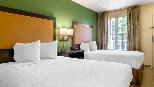 A hotel room with two beds, a nightstand with a lamp, a phone, and a window with curtains. - Extended Stay America Select Suites Raleigh RDU Airport (Morrisville)