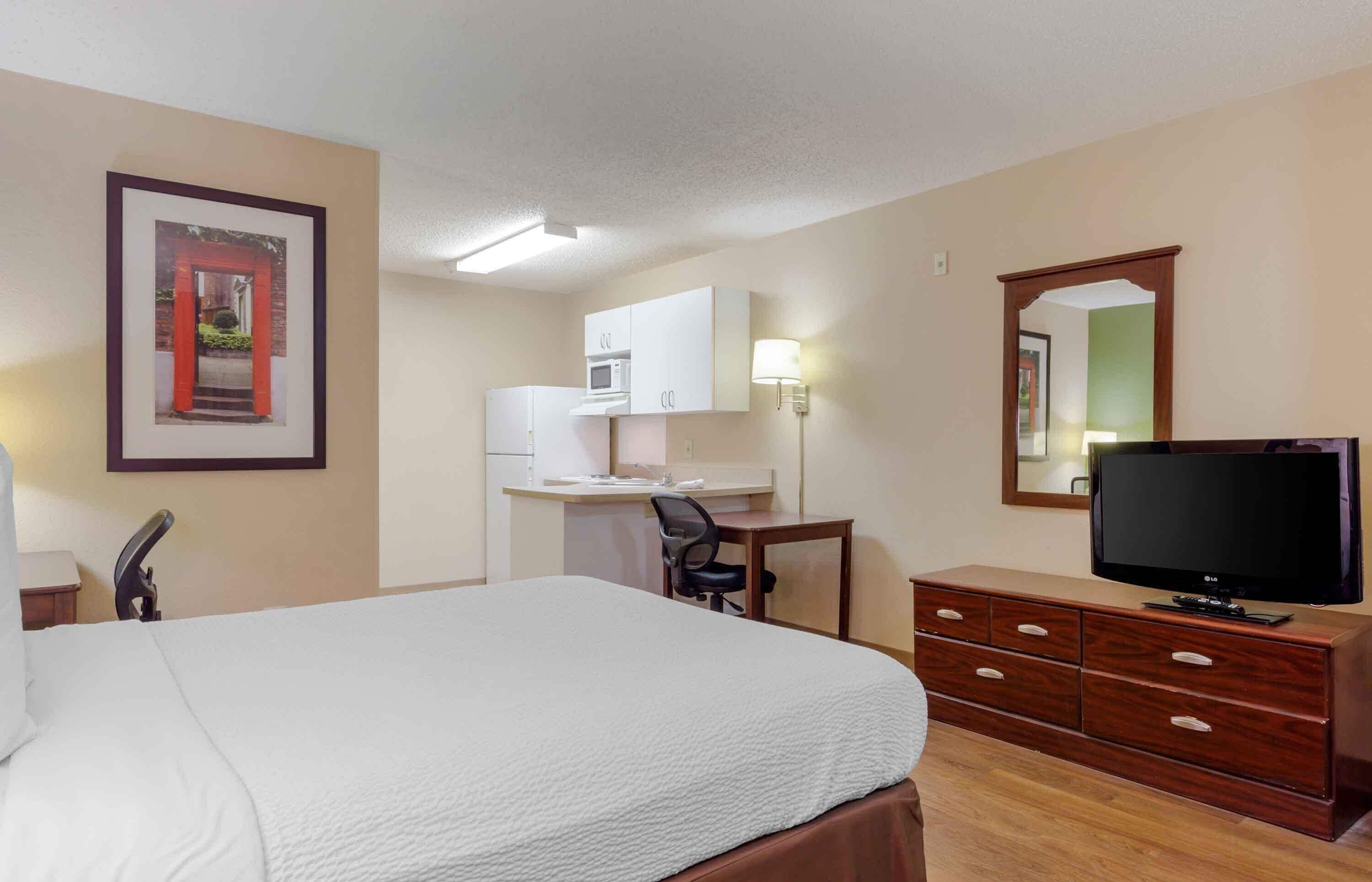 A hotel room with a bed, a desk with a chair, a television, a dresser, a microwave, and a framed picture on the wall.