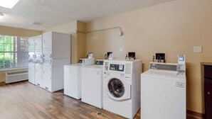 A laundry room with multiple washing machines and dryers, a ventilation system, and a window with a view of greenery. - Extended Stay America Select Suites Raleigh RDU Airport (Morrisville)