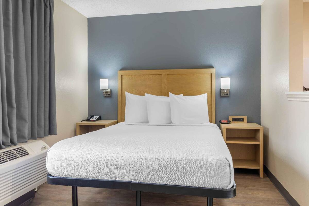 A hotel room with a bed, bedside tables, a phone, a clock, and a wall-mounted light fixture.