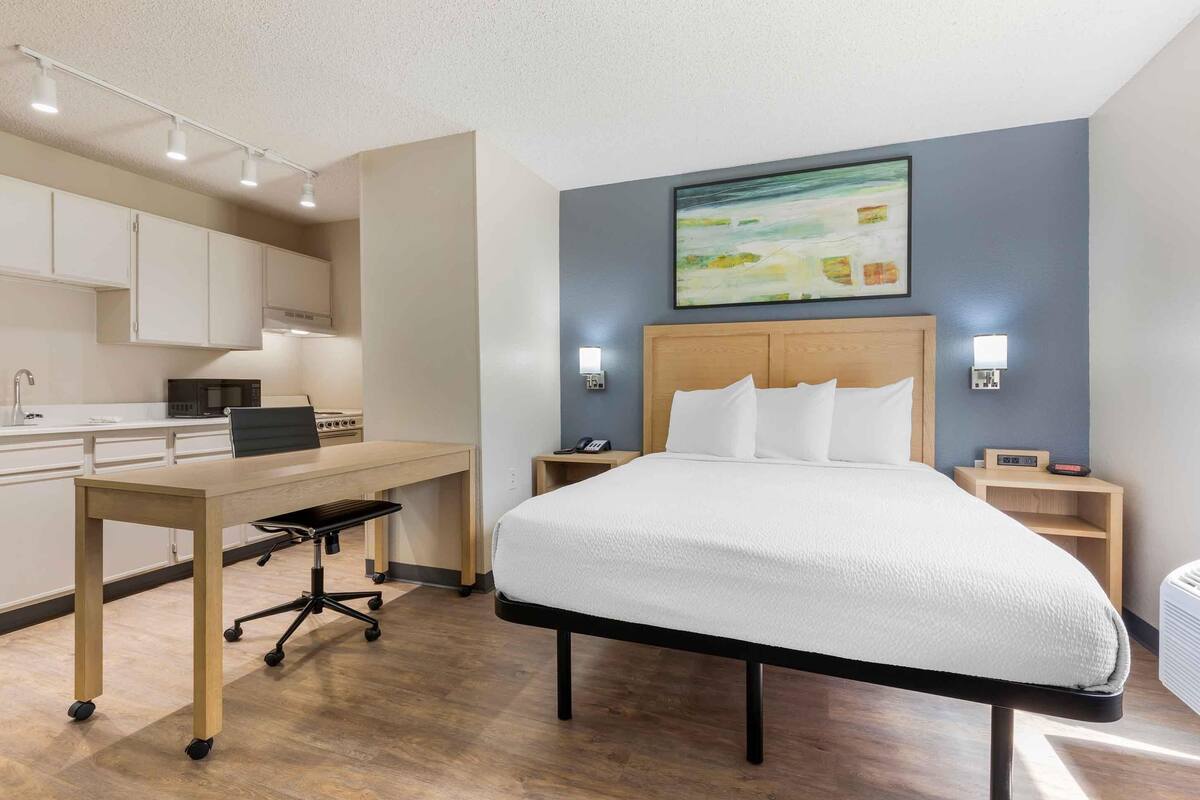 A hotel room with a bed, a desk, a chair, a microwave, and a painting on the wall.