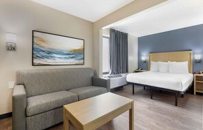 A hotel room with a gray sofa, a wooden coffee table, a bed with white bedding, and a painting on the wall. - Extended Stay America Suites Arlington Six Flags (Arlington)