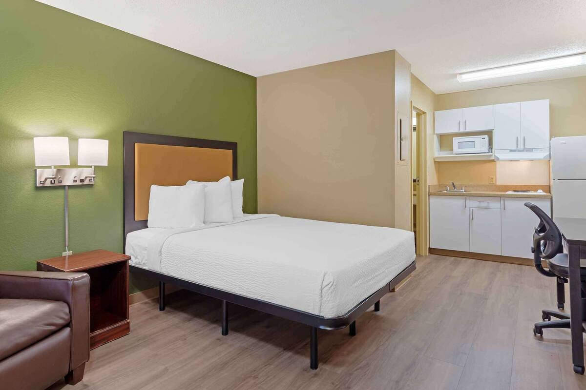 A hotel room with a bed, a desk, a chair, a nightstand, a lamp, and a kitchenette.