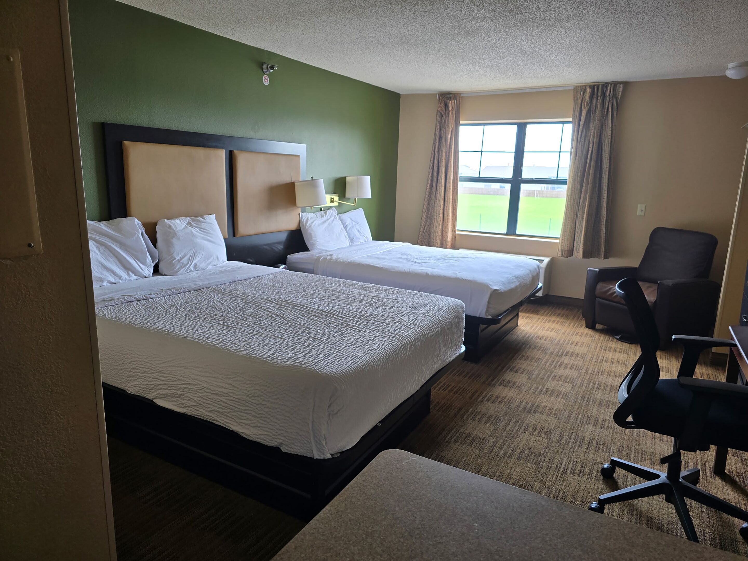 Une chambre d’hôtel avec deux lits, un bureau, une chaise, une fenêtre avec des rideaux et un mur vert.