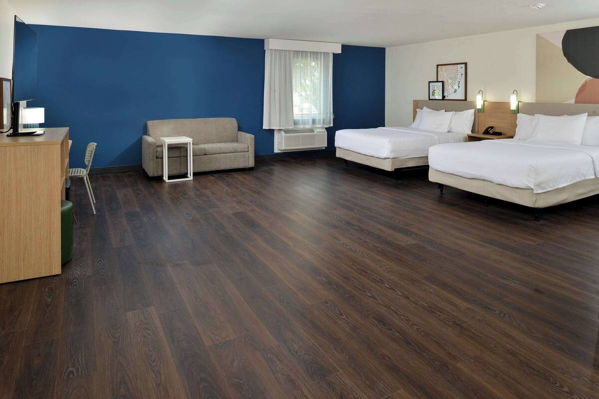 A hotel room with a blue accent wall, a wooden desk with a lamp, a sofa, two single beds, and a small table.